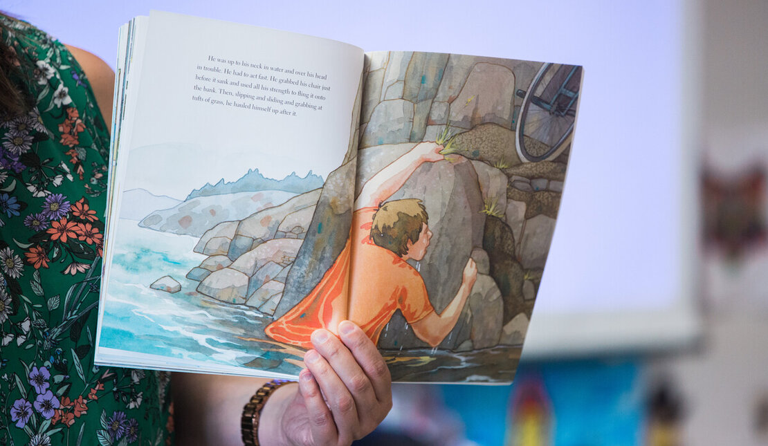 A colourful children's book opened to a page of a child trying to get out of water. His wheelchair is nearby on a rock.