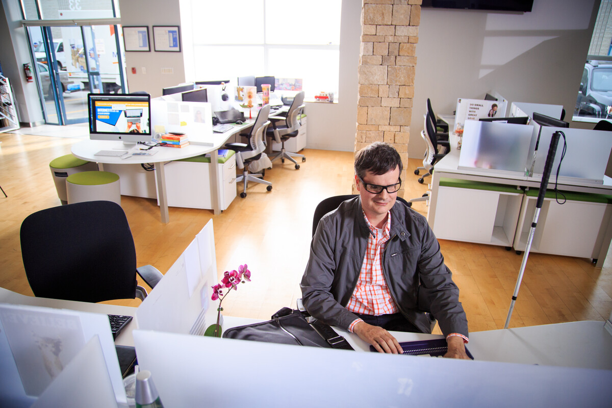 Person seated at a desk in a bright, modern office with workstations, computers, and a white cane leaning nearby.