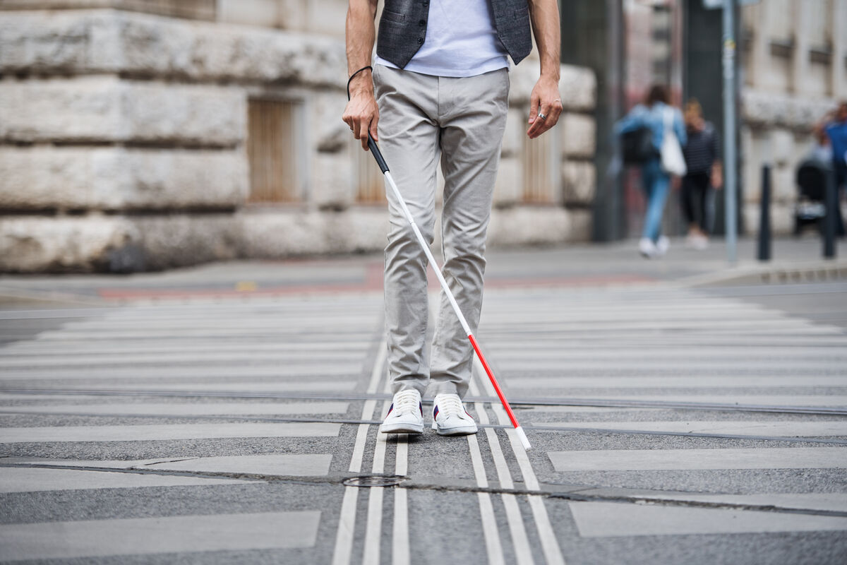 Person using a white cane walks across a city crosswalk with tactile guiding lines embedded in the pavement.