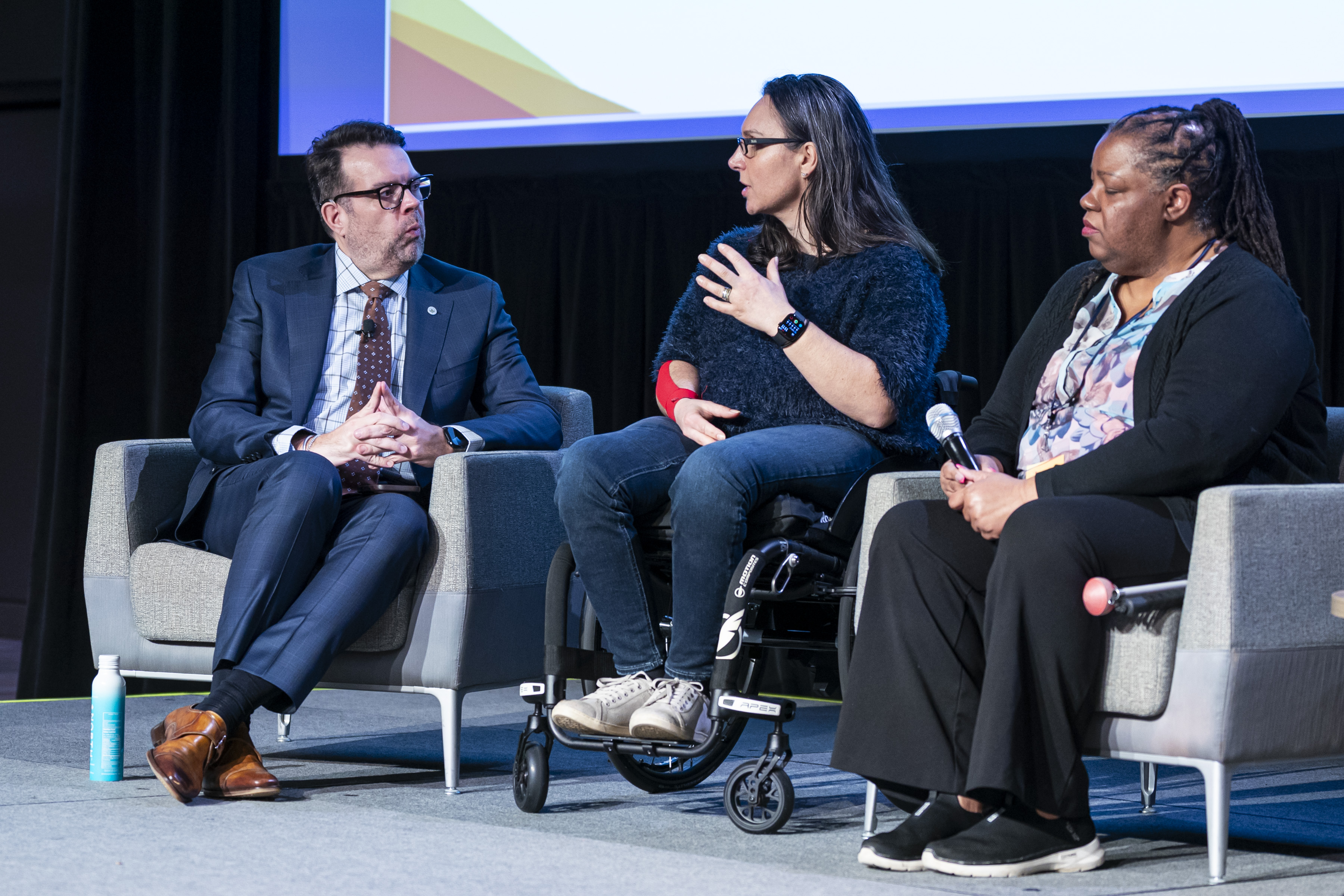 Three individuals seated on stage. The woman in the center uses a wheelchair and gestures while speaking. The person on the right holds a microphone, and the person on the left is dressed in a suit A large screen with colorful graphics is visible in the background.