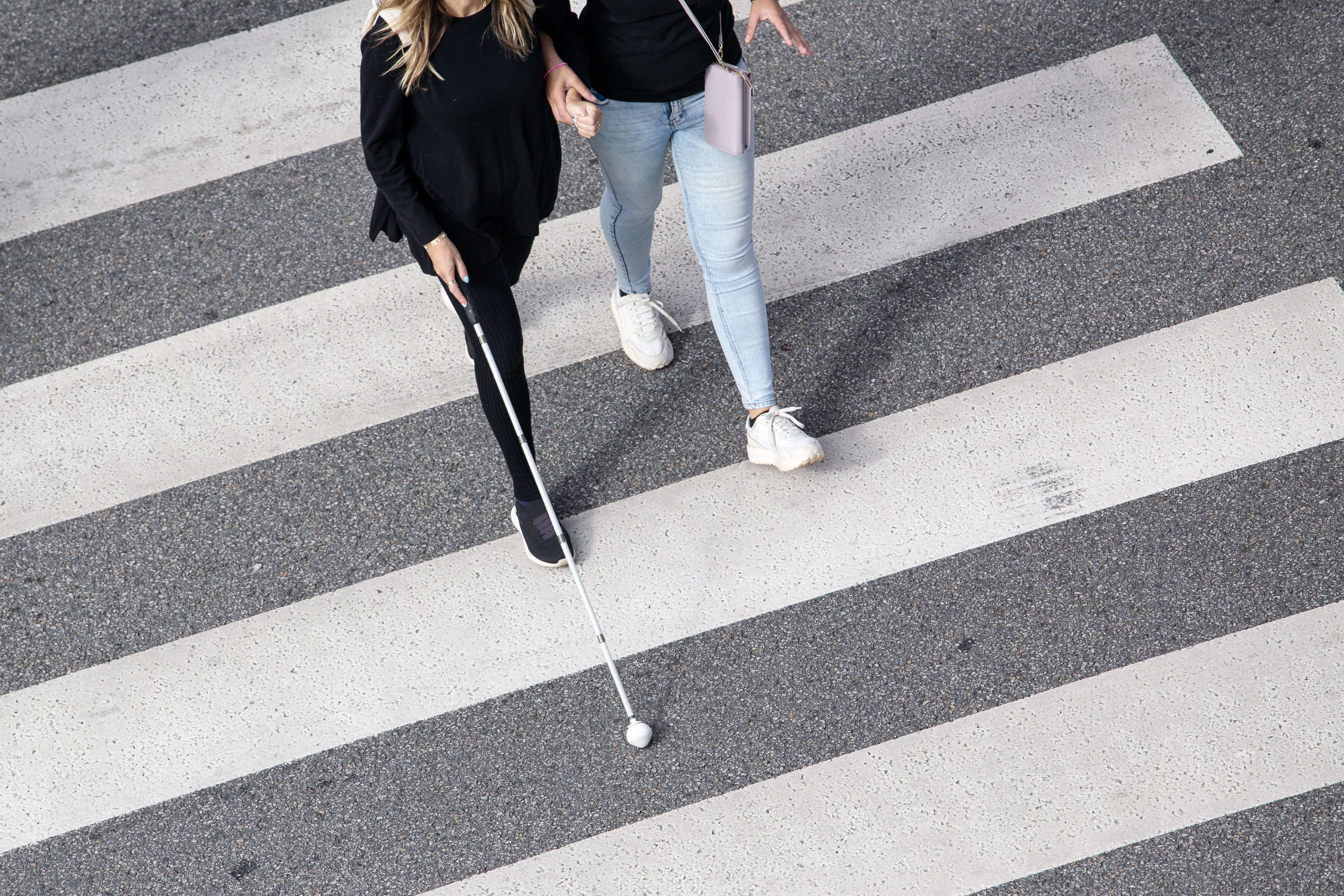 Two people cross a pedestrian crosswalk together, one using a white cane for navigation.