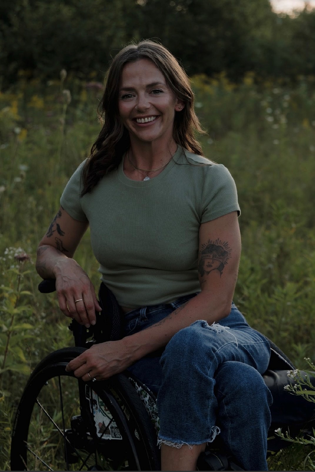 A woman sitting in a wheelchair outdoors in a grassy field, wearing a green shirt and jeans with visible arm tattoos.