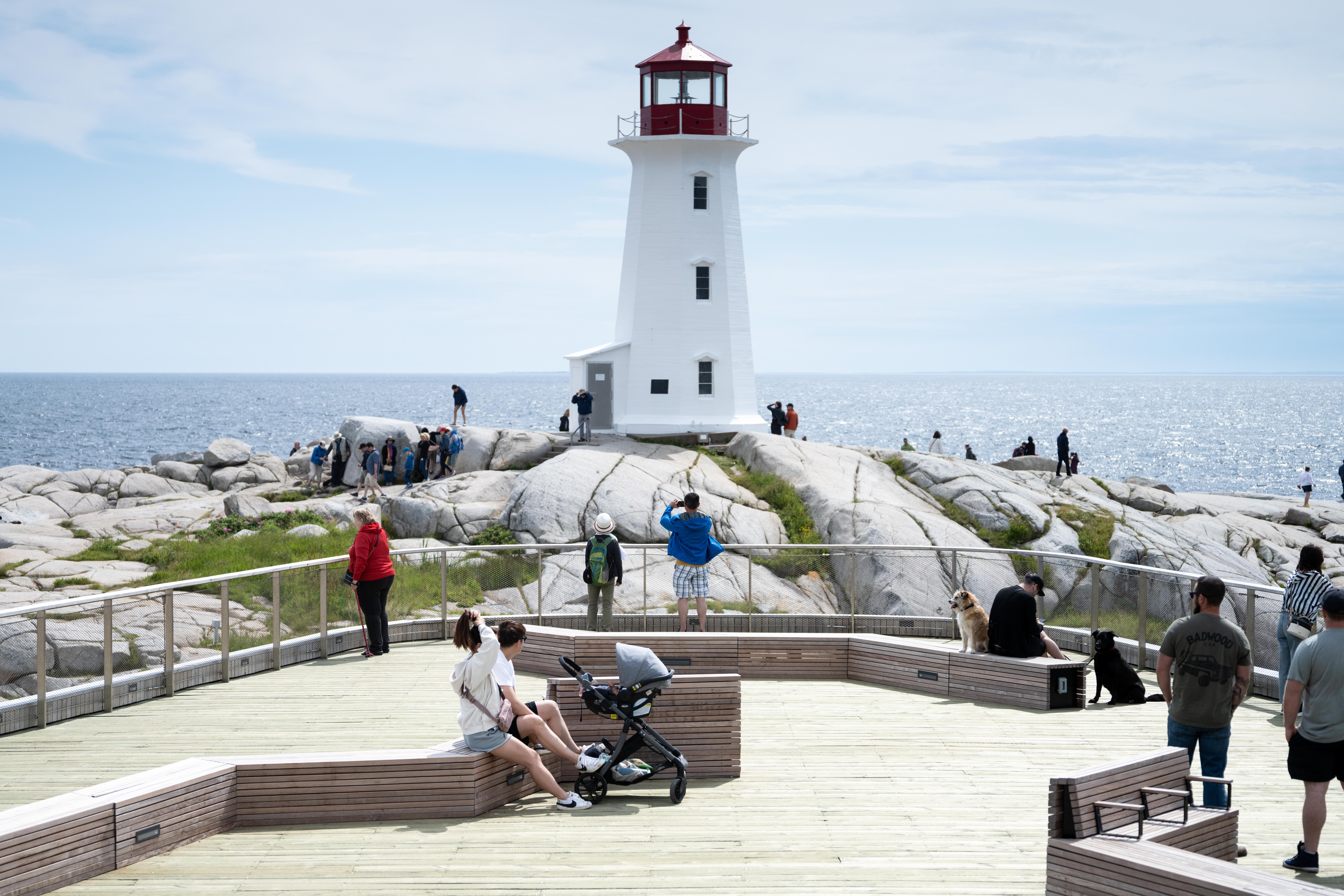 Front of the  Peggy's Cove viewing deck. There are many people using the viewing deck, which is in close proximity to the lighthouse. The lighthouse is white and red and is built on large rocks overlooks the ocean