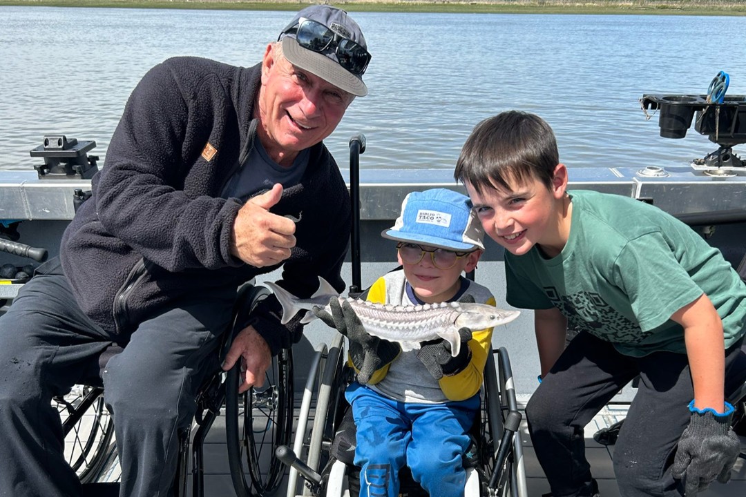 Two children and one adult pose on a dock with a scenic lake and mountains in the background. The adult is and one child are using wheelchairs, and the child is holding a small fish. Accessible fishing accessible equipment is visible on the dock.