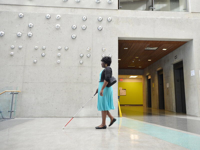 Woman in blue dress using probing cane walks in front of concrete wall with large braille 