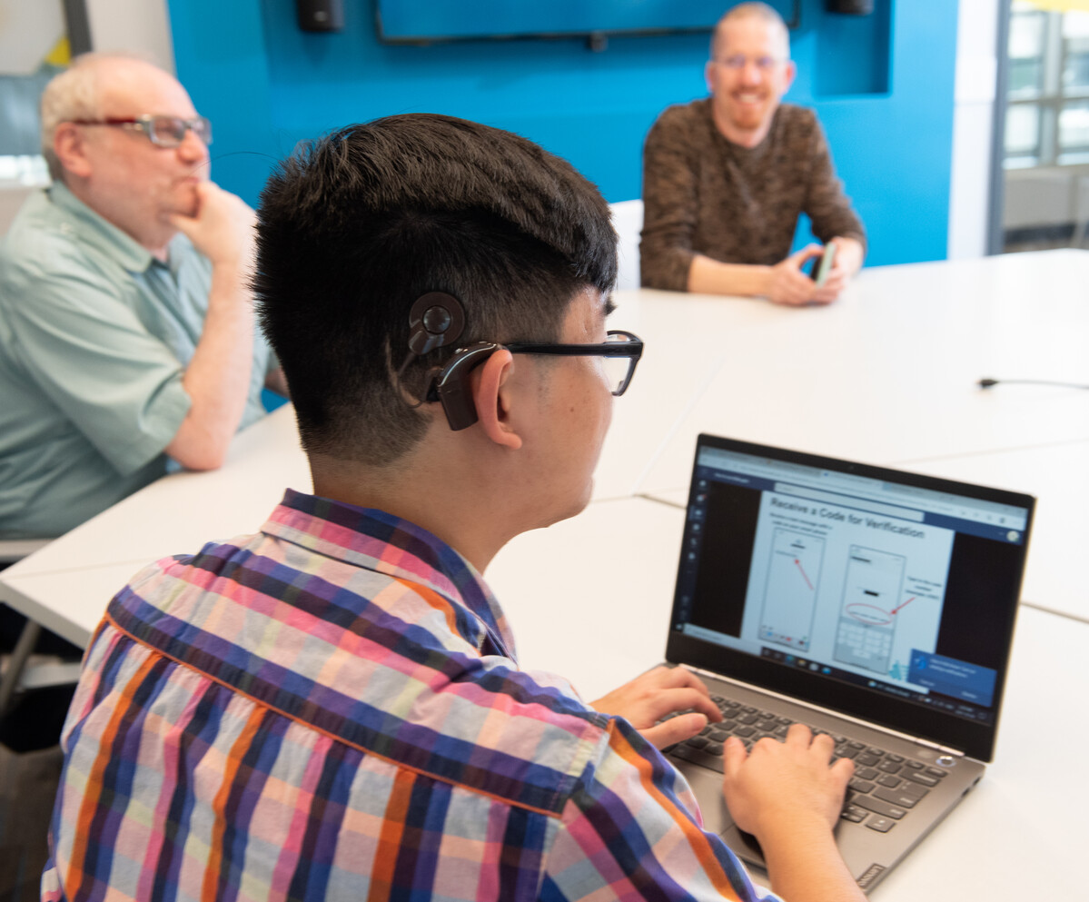 three people at a conference table, one using a hearing aid, using a laptop