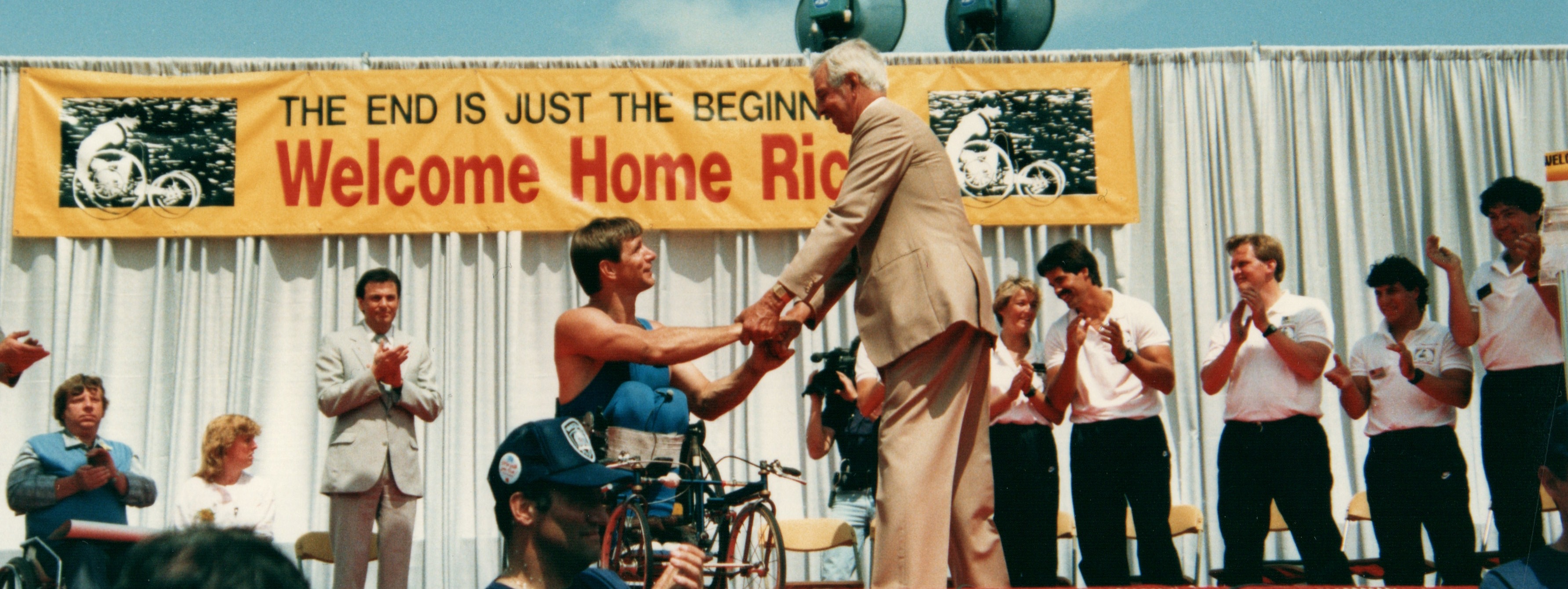 Rick Hansen shakes hands with a middle-aged man on stage while people clap, cheer, and welcome him home after the Tour.