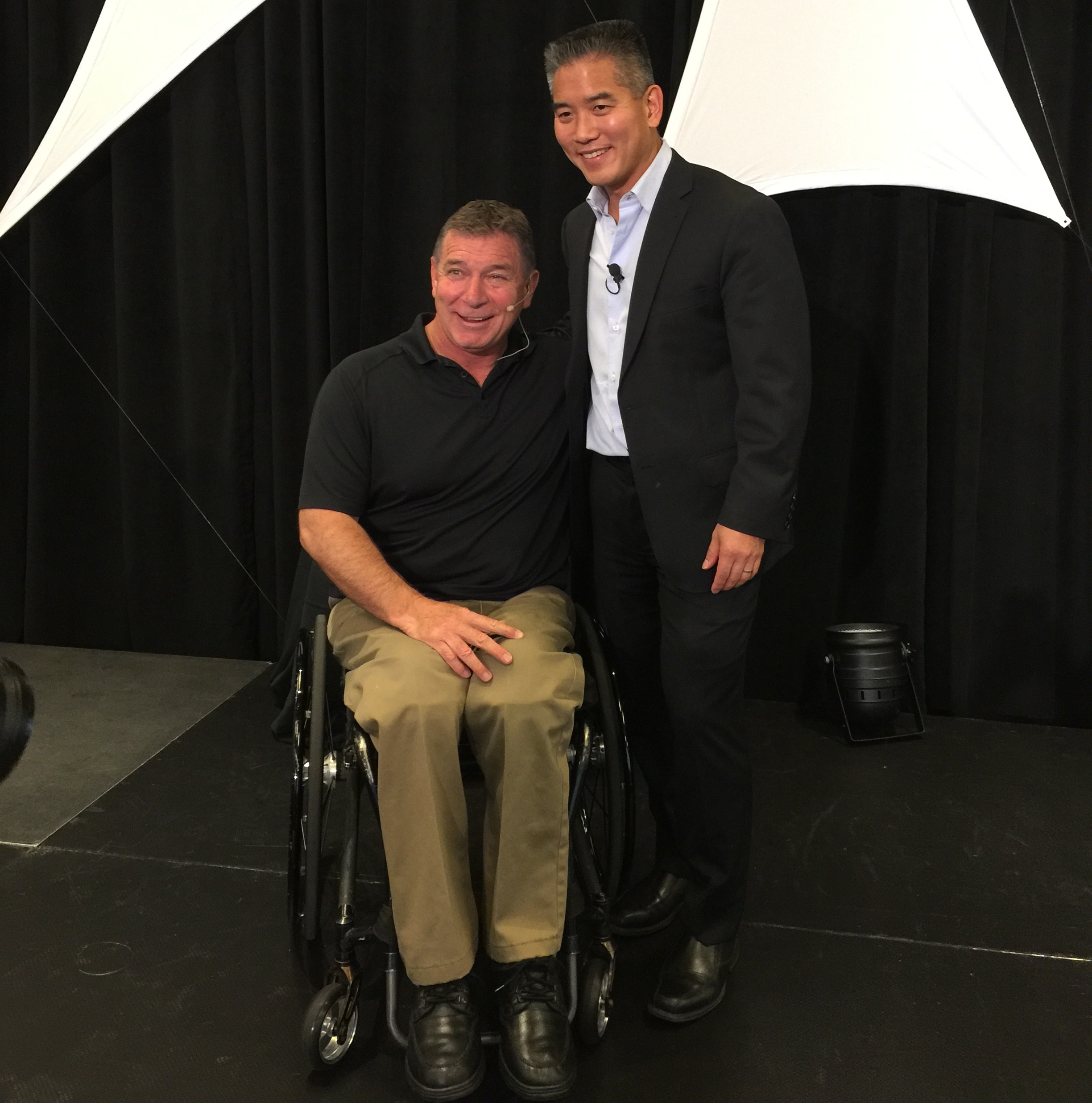 Rick Hansen stands beside Dr. Kwon for a picture on the stage. Rick is wearing a black t-shirt with beige pants and Dr. Kwon is wearing a black suit with a light blue button-up shirt.