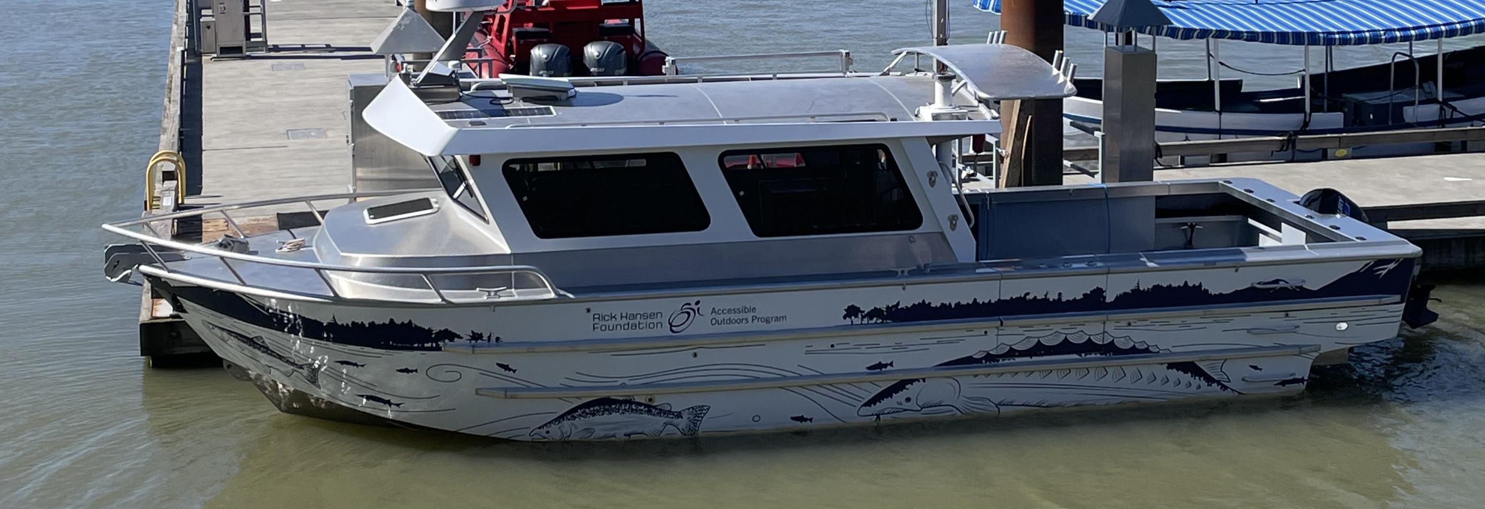 A small silver boat with the RHF Accessible Outdoors Program labelled on the side. It is stationed beside the dock.