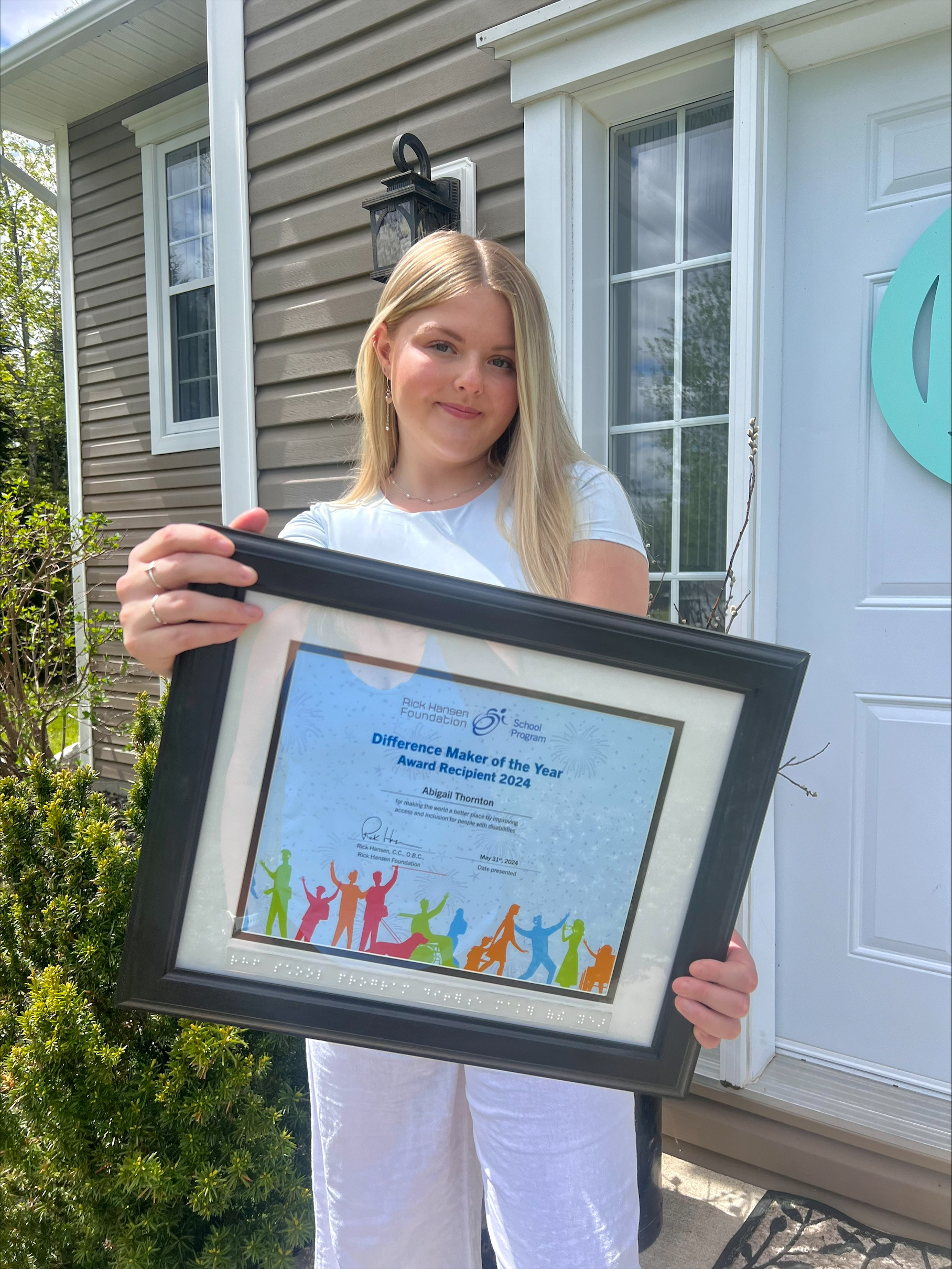 Abigail Thornton stands in front of a house holding her framed Difference Maker of the Year Award 