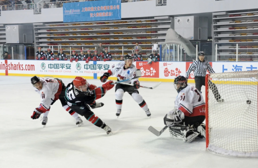 Photo of four hockey players, including Adam Taylor, playing a game for the China Sharks. In the image, Taylor is shooting and scoring on the opposing goalie. 