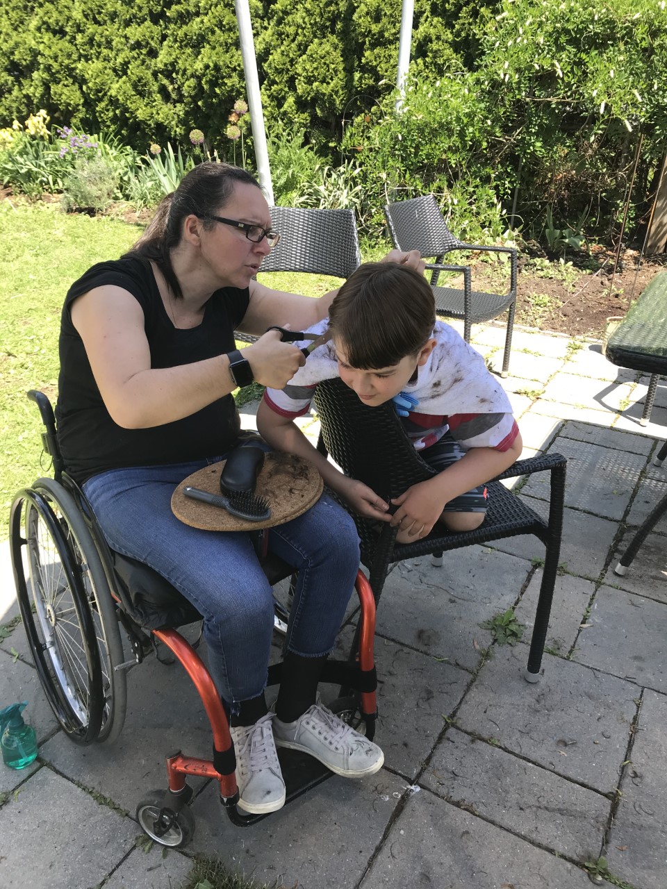 Woman sitting in wheelchair cuts boy's hair outside.