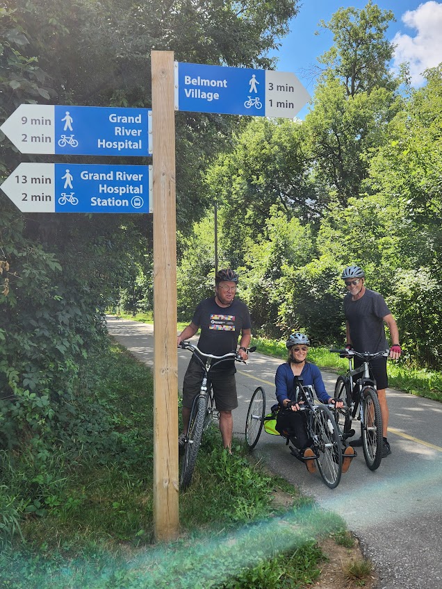 A picture of Cindy with two male friends on their bikes in the park. They are standing on a pathway beside a sign post. Vibrant green trees surround them. 