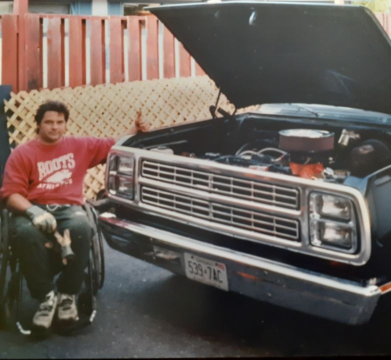 Dave Padovese wears a red long sleeve shirt and poses beside his a black car that he worked on as a mechanic.