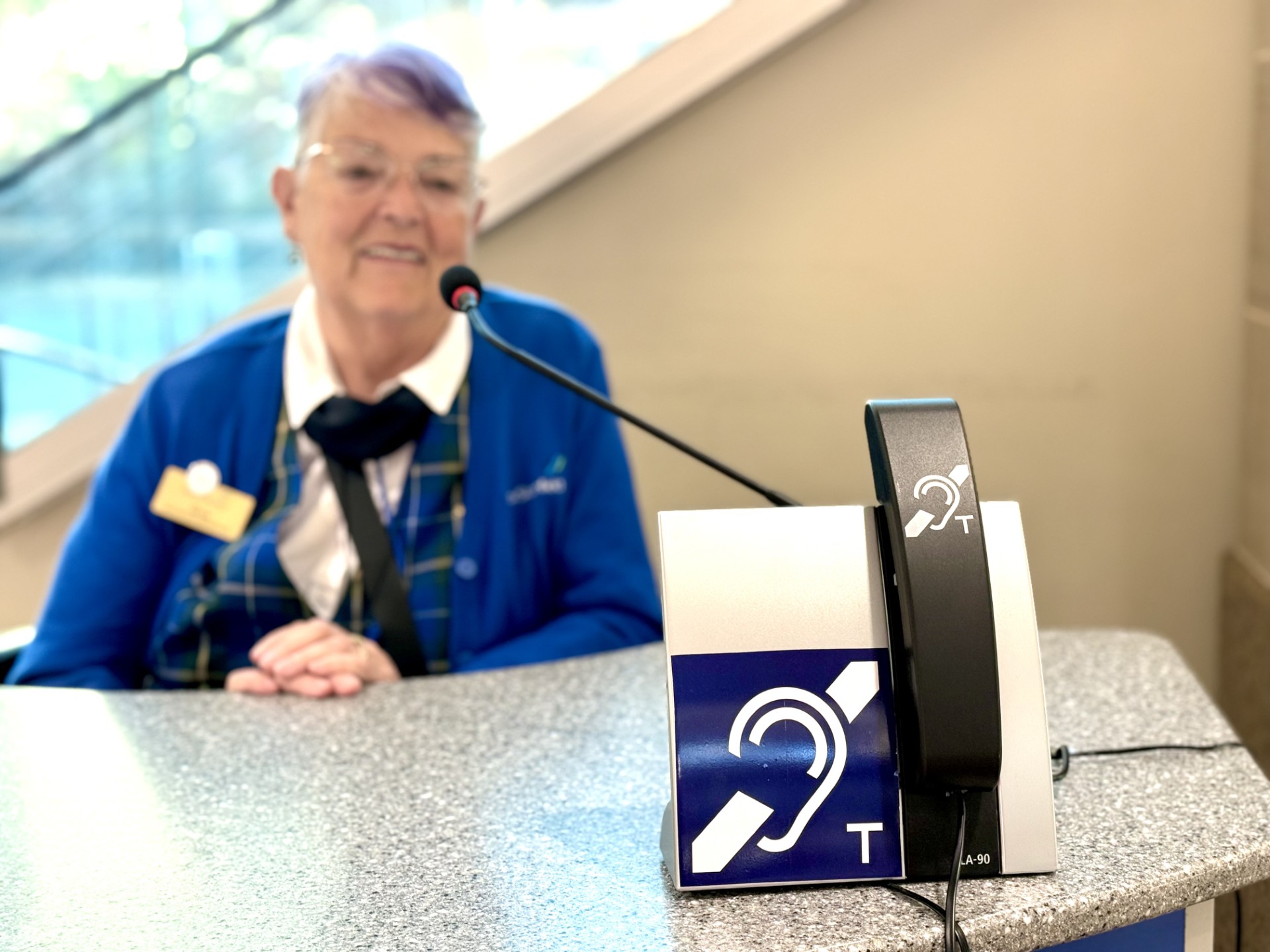 A photo of a hearing loop device that is available for use at a counter in the airport. A staff member smiles behind the counter.