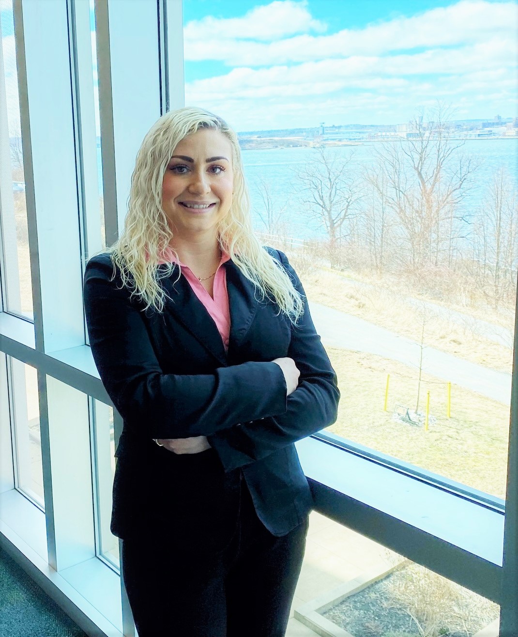 Melissa is wearing a black blazer with a pink button-up shirt underneath and black pants. She has a bright smile and platinum blonde curly hair that runs down past her shoulders.