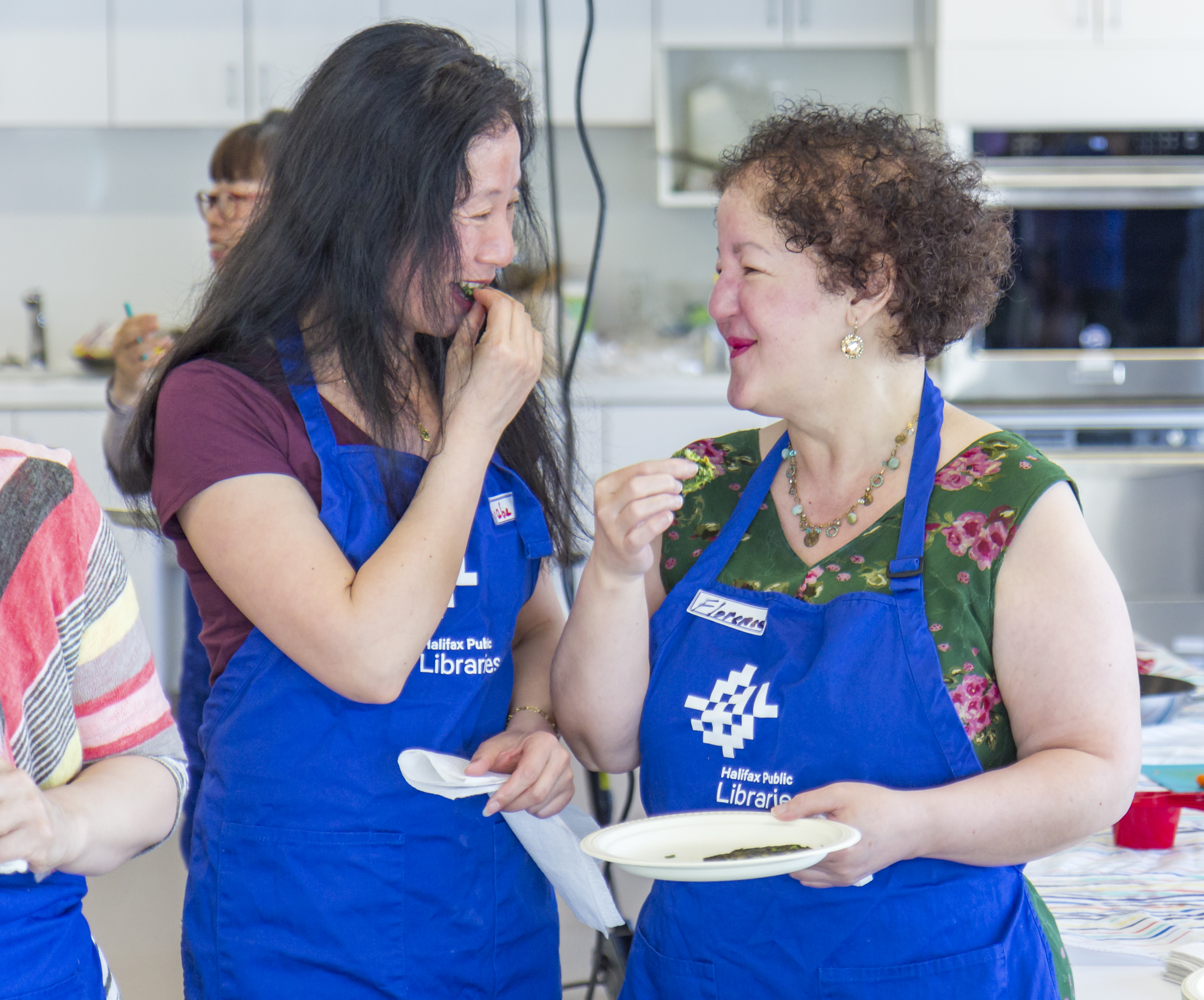 Two women taste food in a kitchen wearing matching aprons