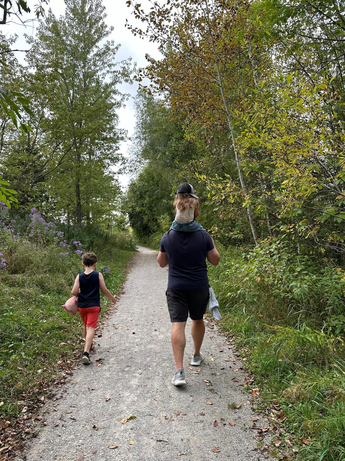 Matt Shaw out on a walk with his son walking on trail beside him and his daughter on his shoulders.