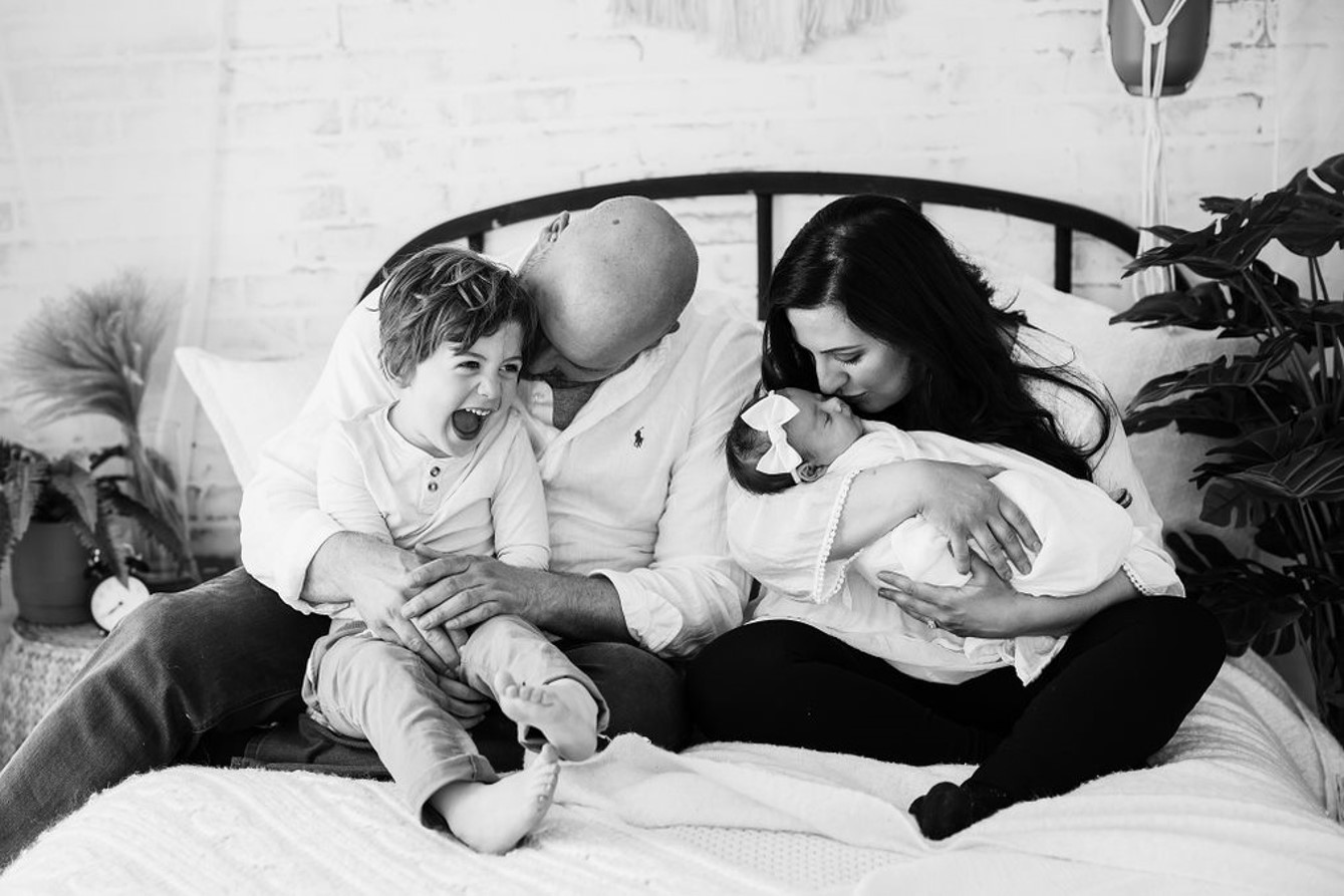 A black and white photo of Matt shaw holding his son while his wife is cradling their daughter. They are all sitting on a bed.