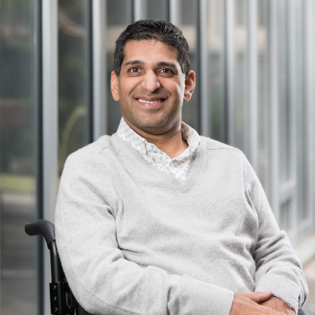 A headshot of Nabeel. He is wearing a white sweater and is smiling and using his wheelchair. 