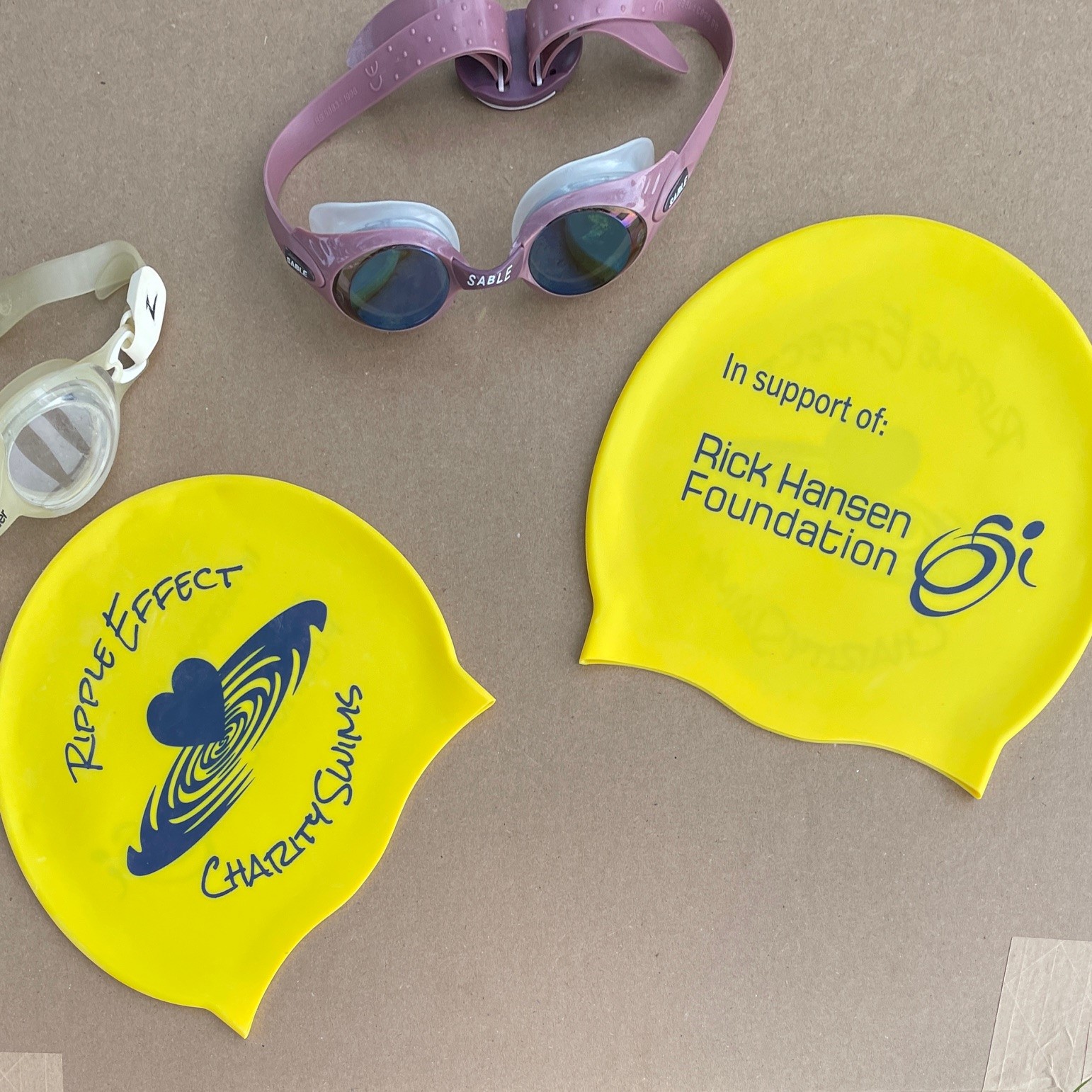 Two yellow swimming caps that have the RHF logo on one side and the Ripple Effect Charity Swims logo on the other. There are two white and purple swimming goggles beside the caps.