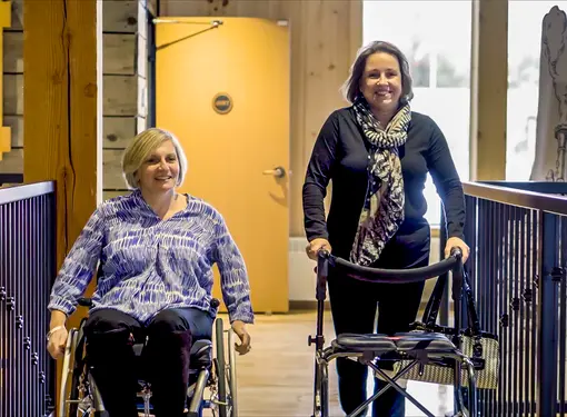 Two women smiling for camera. One uses a wheelchair and one uses a standing frame.