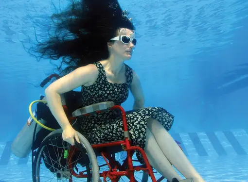 Woman in wheelchair underwater at bottom of pool with goggles 