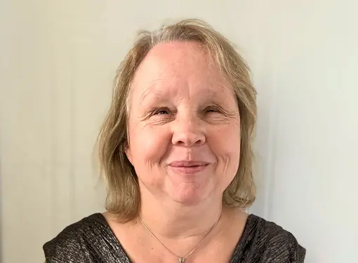 middle aged woman with light colored hair smiles against a plain backdrop, wearing a pendant necklace and a flowy blouse