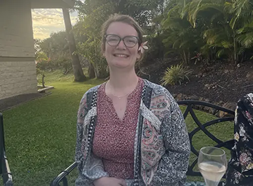 Alana is seated at an outdoor dining table surrounded by blue skies, palm trees, and a building. She is wearing a floral decoration on her hair, glasses, and smiling in a summer floral outfit.