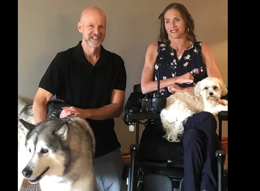 woman with shoulder length brown hari in a wheelchair, smiling with one dog on her lap and another beside her, and a man in a polo shirt kneeling next to them, smiling, with close cropped greying hair