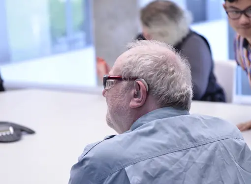 AN elderly man who is wearing glasses and has a hearing aid. He is sitting at a table with others.