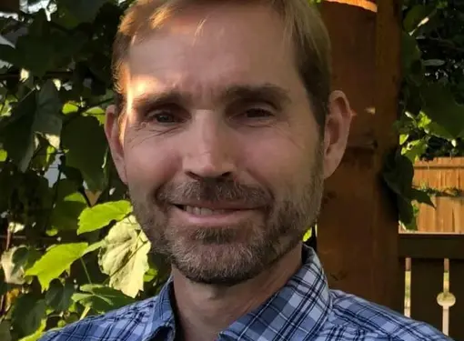 A headshot of Keith outside in front a tree. He is wearing a blue plaid shirt and has short brown hair.