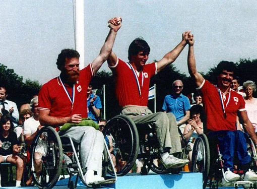 A picture of Rick Hansen and his two colleagues are on a blue pedestal stand holding each other's hands up in the air in celebration for winning in the Paralympic race. The three of them are wearing gold medals and the same red team shirts.