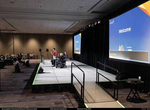 A conference room with a few guest speakers standing on a low-level stage that has a ramp. Behind the stage are two big screen projectors by the wall. The room is lightly dimmed.