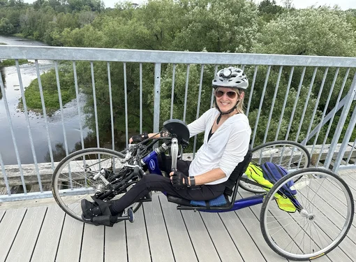 A picture of Cindy who is wearing a helmet, white shirt, and sunglasses. She is pictured on a bridge with the water and trees in the background.