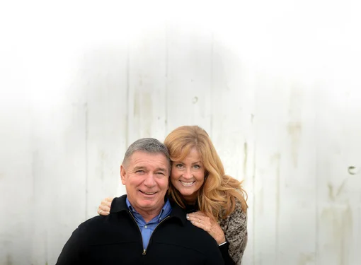 Rick and Amanda Hansen, smiling against a white backdrop, with Amanda hugging the Rick lovingly from behind.