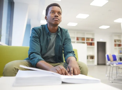 A young male teen is reading a textbook in braille at a library. He is wearing a green long sleeve shirt on top of a gray t-shirt and beige pants.  