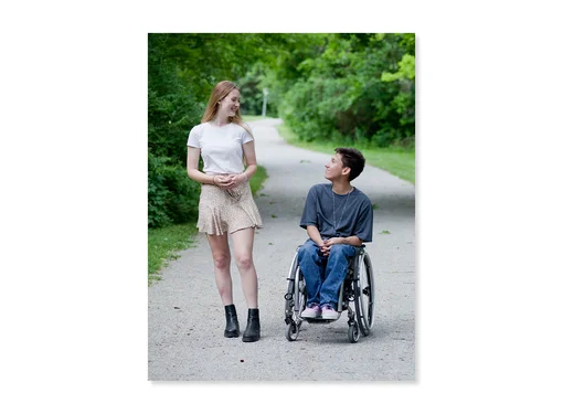 Two friends on a path in a park, smiling at each other. One is standing and one is sitting in their wheelchair. 