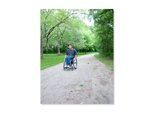 Teenager using a wheelchair on a path surrounded by greenery. 