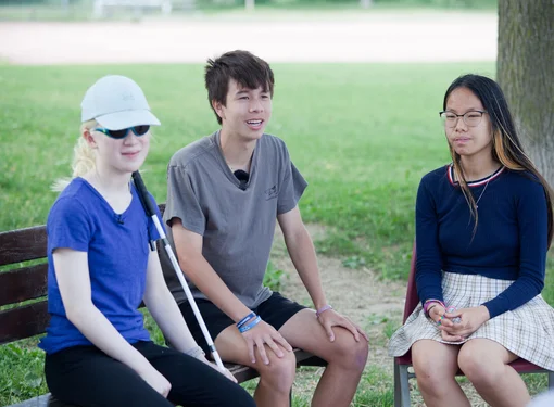 Three students sitting on a bench together. On the left is a female student with a blue shirt and using a white cane. The middle student is in a grey shirt and the final student in a blue sweater. 