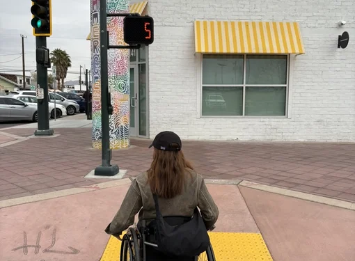 A photo of Emily from behind rolling in her wheelchair over a curb cut. 