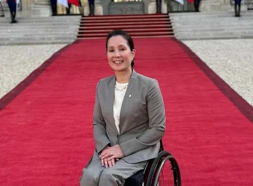 Photo of Miki Matheson in a tan suit using her wheelchair on a long red carpet leading to a building