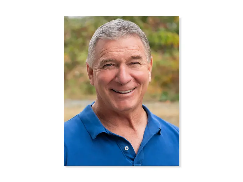 Headshot of Rick Hansen. He is smiling, wearing a blue golf shirt. In the background there are trees and greenery.