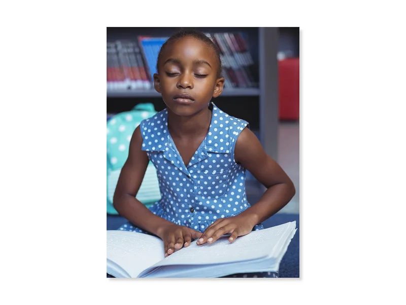 Une jeune fille à la peau foncée, vêtue d’une robe à pois bleus et blancs, lit un livre en braille.