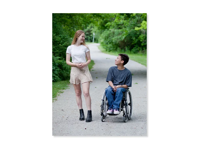 Two friends on a path in a park, smiling at each other. One is standing and one is sitting in their wheelchair. 