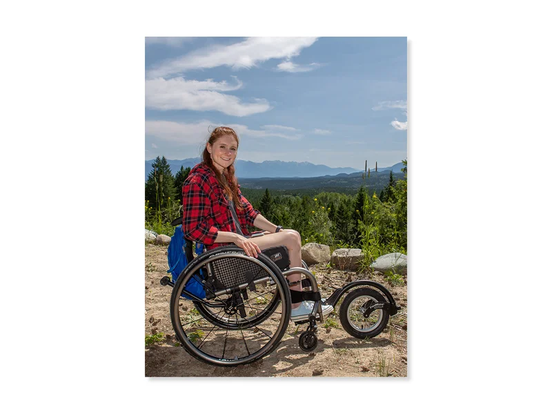 Une jeune femme en chemise à carreaux est assise dans son fauteuil roulant au sommet d’une montagne. Son fauteuil roulant est équipé d’un accessoire tout-terrain.