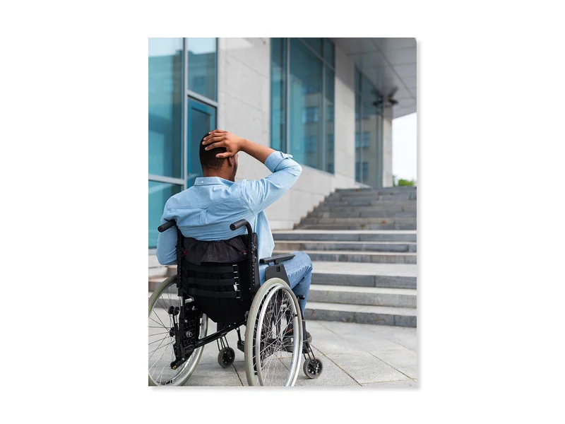 Man in a wheelchair, looking at a set of stairs in front of him. The man has one hand on his head expressing uncertainty of how to surmount the barrier. 