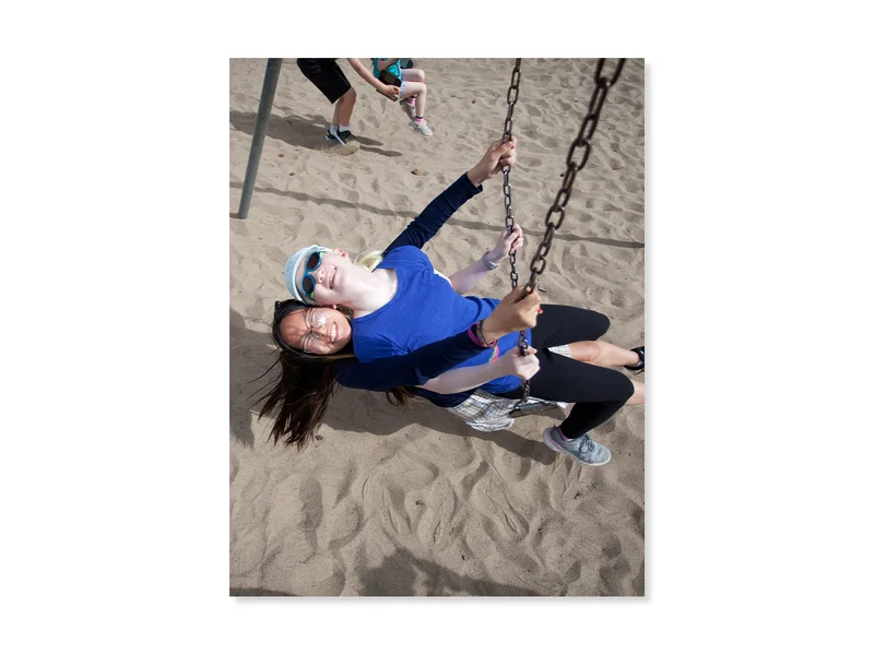 At a playground, two sisters smiling big and swinging together on the same swing. 