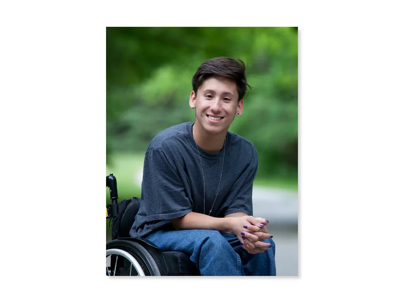 Teenager with short, dark brown hair smiles at the camera. They are using a wheelchair and wearing blue jeans, a grey t-shirt and their nails are painted purple.