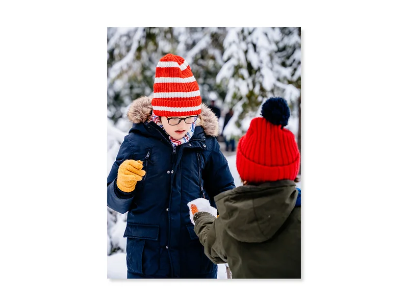 Two children outdoors in their snowsuits, one child is showing the other a snowball. 